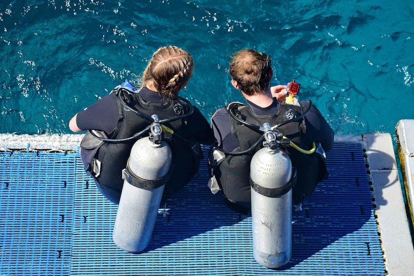 Two divers with scuba tanks seated on a boat deck by a clear blue sea. 3