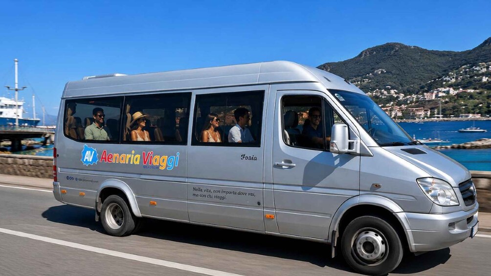 A silver van with 'Aenaria Viaggi' branding, driving on a road with a coastal backdrop. 2