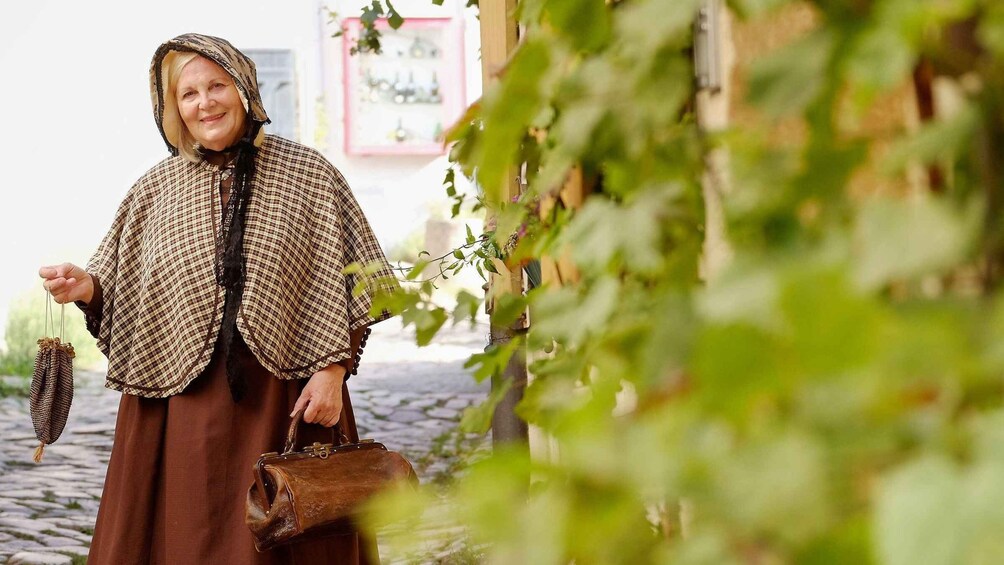 A person holding a small woven bag and a rolled-up item, standing in a cobblestone area with greenery and a building in the background. 1