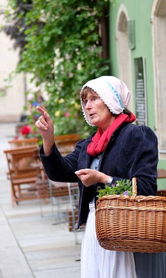 A person gesturing with one hand, wearing a dark jacket, white skirt, and a red scarf, holding a wicker basket with greenery. 5