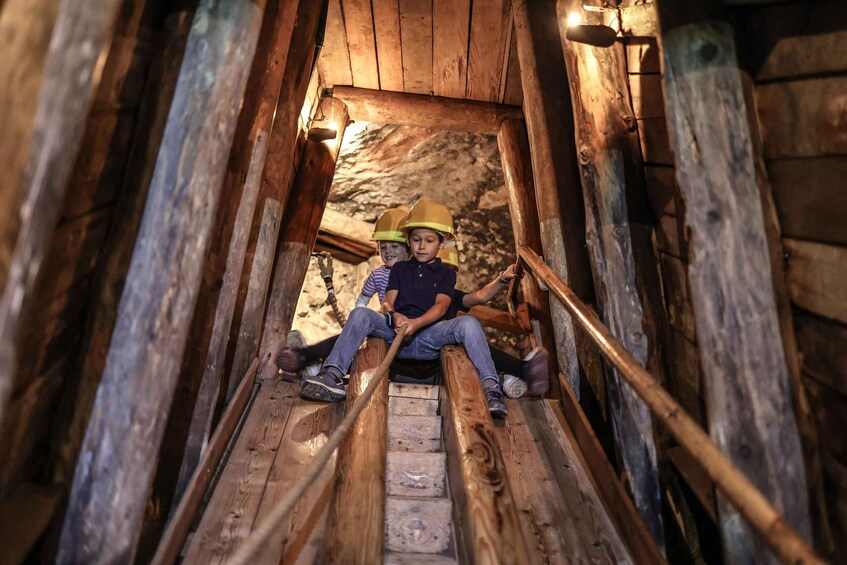 Two individuals in hard hats sitting on wooden stairs in a wooden structure with a cave-like background. 2