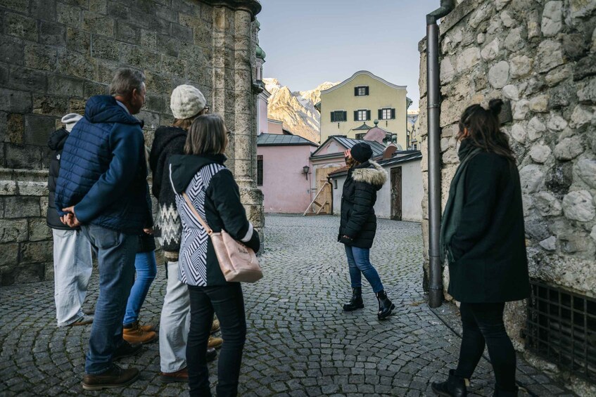 A cobblestone street with historic stone buildings, a mountain backdrop, and people walking. 1