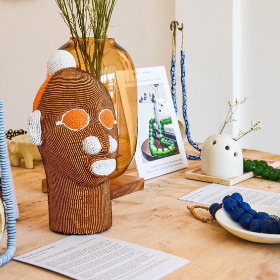 A crocheted sculpture, a vase with green plants, a framed picture, a white ceramic pot, and a plate with blue beads. 1
