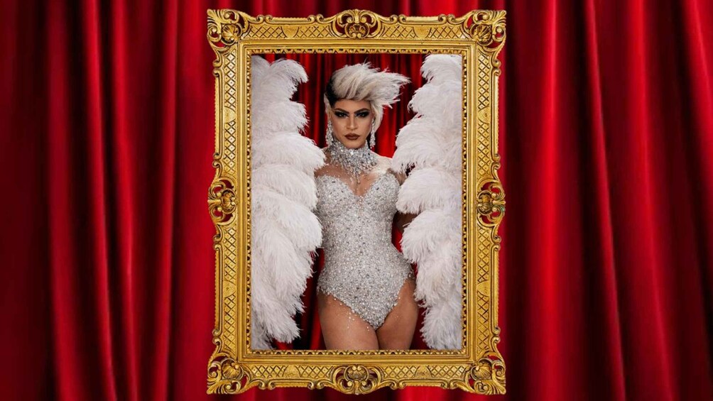 A person in a sparkling, sequined outfit with white feathered wings, standing in front of a red curtain backdrop. 2