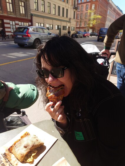 A person eating a sandwich at an outdoor table in an urban setting. 5