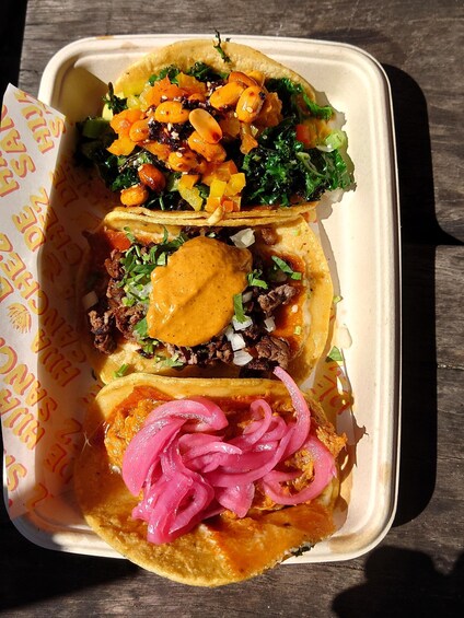 Three tacos in a takeout container with toppings like lettuce, tomatoes, onions, and pickled onions. 2