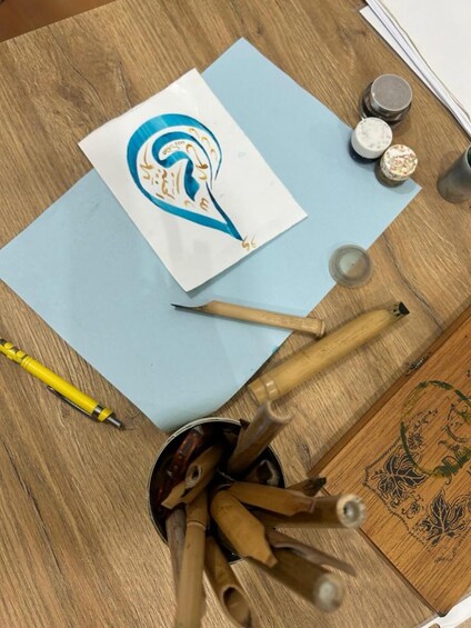A wooden table with a blue sheet of paper, a yellow pen, a container with bamboo brushes, and a small jar of white powder. 2