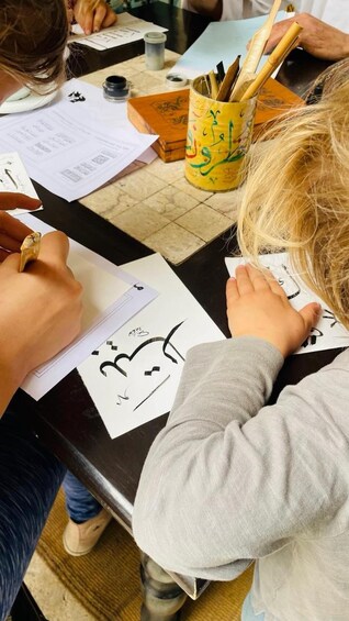 A person writing on paper with a pen, a yellow cup with writing utensils, and a notebook on a table. 4