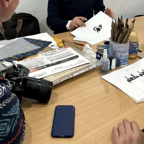 A person holding a white card with a black symbol, a camera, a smartphone, and various stationery items on a table. 3