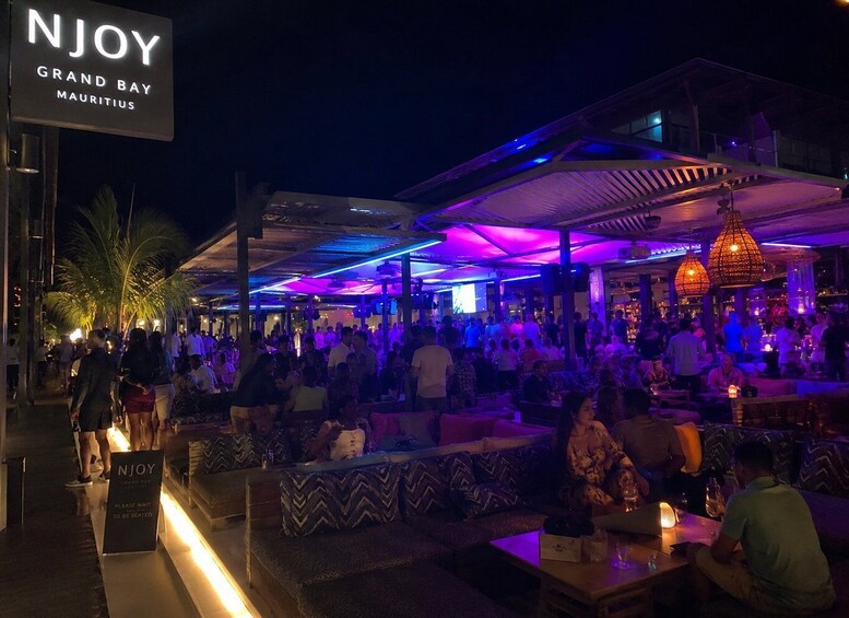 A crowded outdoor bar with neon lights and a sign reading 'NJOY GRAND BAY MAURITIUS.' 2