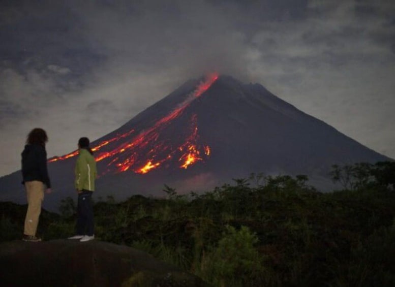 Two people observing a volcano with lava flowing down its side. 5
