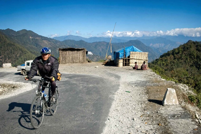 A cyclist on a paved road with mountains in the background, a small hut, and a blue tarp-covered structure. 2