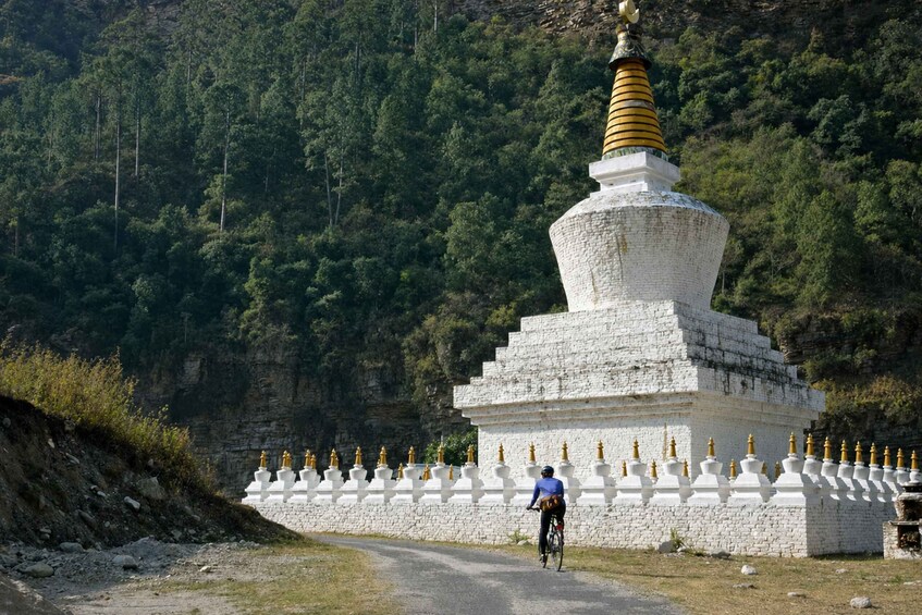 A person riding a bicycle past a white stupa with a golden spire, surrounded by a white fence and set against a forested hillside. 1