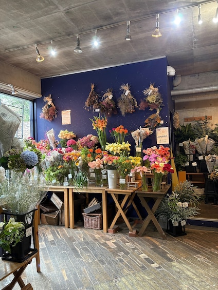 A flower shop with various flowers in pots and vases, a blue wall, and a concrete ceiling. 2