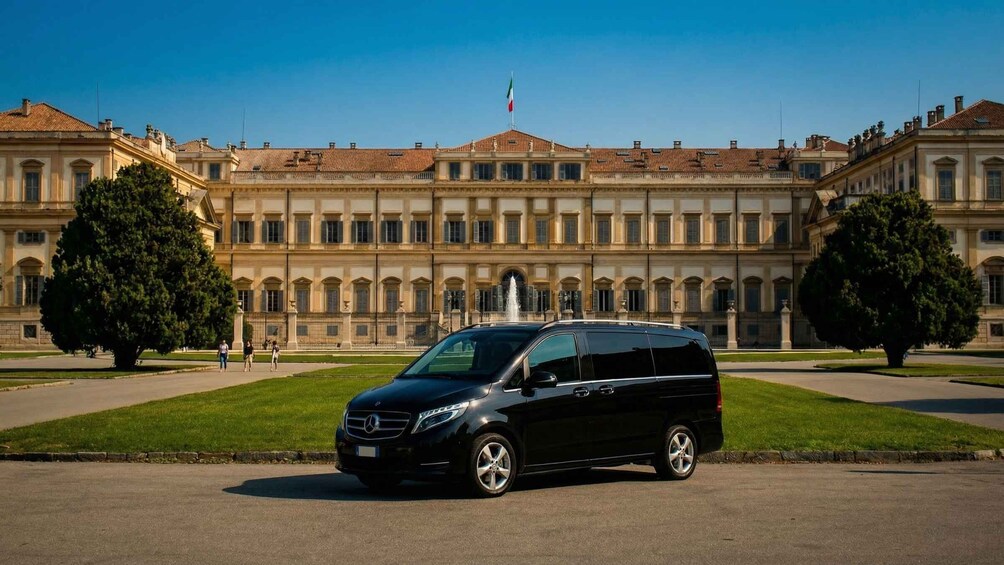 Musta Mercedes-pakettiauto oli pysäköitynä suuren, klassisen rakennuksen eteen, ja rakennuksen keskellä oli suihkulähde. 5