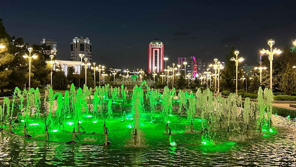 A nighttime fountain display with green lighting, surrounded by street lamps and buildings in the background. 2