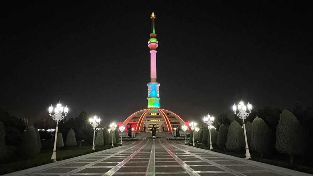 A brightly lit tower with a pink and green color scheme, surrounded by a symmetrical arrangement of lamp posts and trimmed hedges. 4