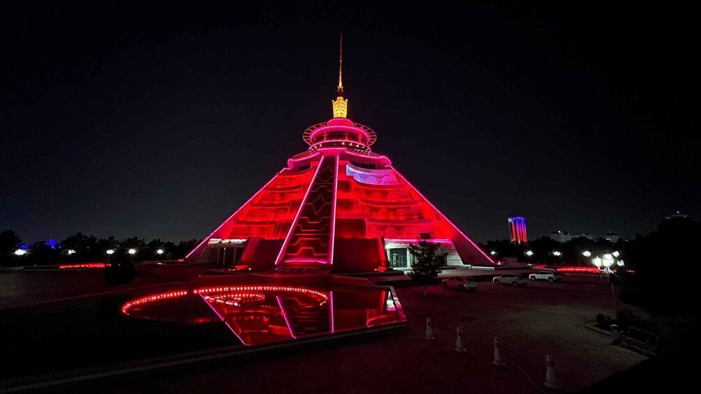 A pyramid-shaped building with a spire, illuminated in red, surrounded by a reflective pool and traffic cones. 1