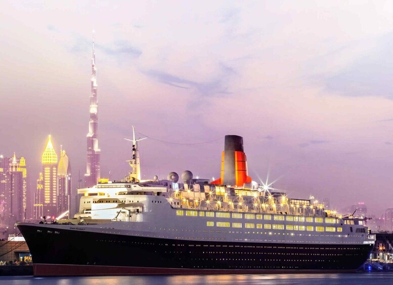 A large cruise ship with multiple decks and a prominent funnel, docked in a city with high-rise buildings in the background. 1
