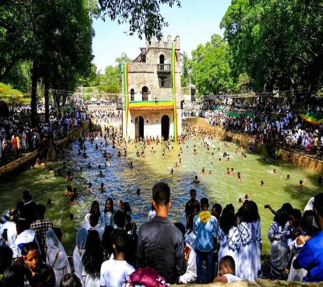A large crowd gathered around a historic stone structure with a tower, surrounded by a water-filled moat. 1