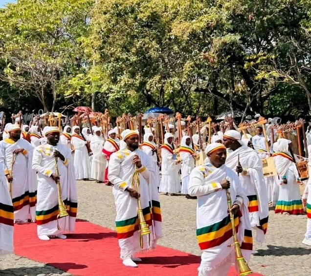 A group of people in traditional attire with staffs, standing on a red carpet in an outdoor setting. 2