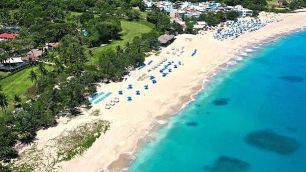 A beach with clear turquoise waters, a row of blue umbrellas, and a line of houses in the background. 5