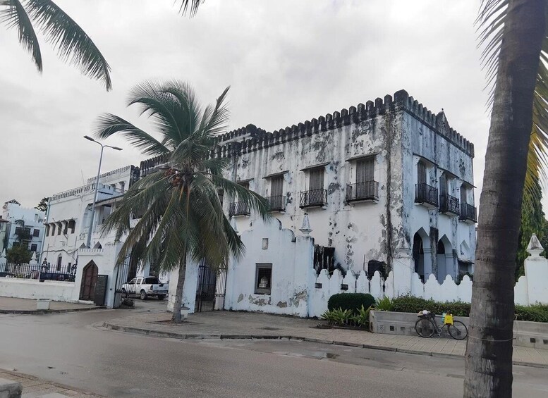 A weathered white building with arched windows and balconies, a palm tree in the foreground, and a bicycle parked on the sidewalk. 1