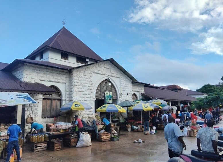 A bustling outdoor market with vendors and shoppers, a stone building with a peaked roof, and umbrellas providing shade. 4