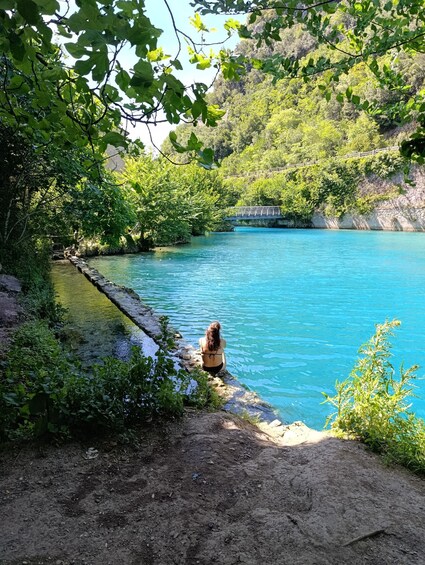 A person sitting by a clear blue river, surrounded by lush greenery and a stone bridge in the background. 2