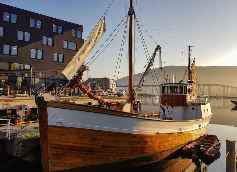 A wooden boat docked at a marina with a building and a bridge in the background. 2