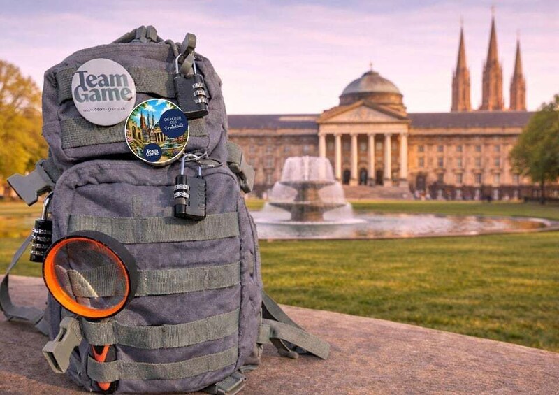 A backpack with 'Team Game' stickers, a camera, and a strap. In the background, there is a building with a dome and spires, and a fountain. 1