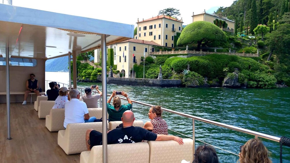 Passengers on a boat with a view of a lake, buildings, and a well-maintained garden. 1