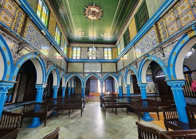 An ornate hall with blue and gold patterned walls, wooden benches, and a central chandelier. 2