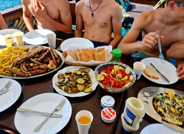 A group of people dining on a boat with plates of food including sausages, fries, bread, and vegetables. 5