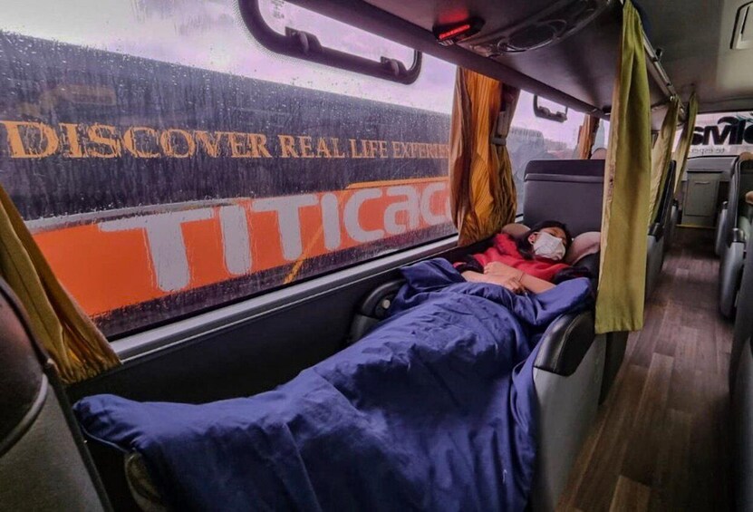 A person lying on a bus seat covered with a blue blanket, with a window displaying the text 'DISCOVER REAL LIFE EXPERIENCES' and 'TIC TACO'. 5