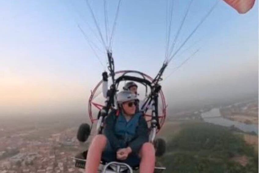 A person in a paragliding harness with a wing attached, flying over a landscape with a river and buildings. 3