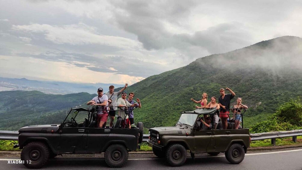 A group of people on a jeep and a pickup truck on a road with a mountainous landscape in the background. 1