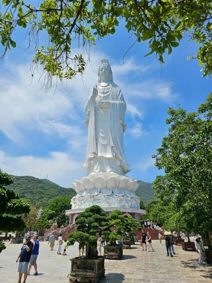 A large white statue of a robed figure with a lotus base, surrounded by visitors and trees. 4
