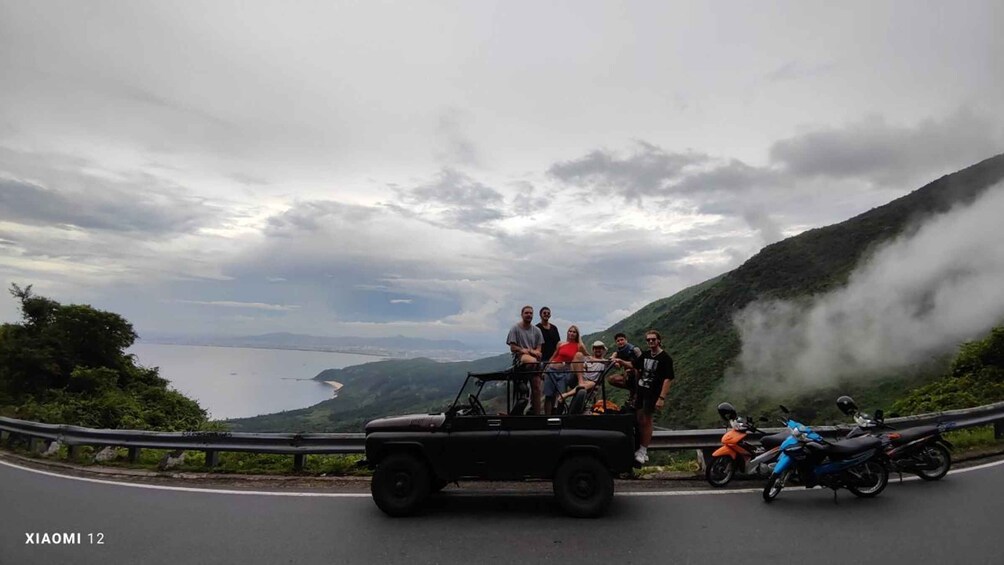 A group of people on a vehicle and motorcycles on a road with a mountainous landscape and a body of water in the distance. 2