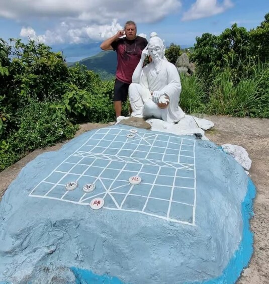 A person standing next to a large, painted board with a grid and symbols, set against a natural landscape with trees and a mountain in the background. 4