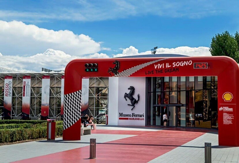 A red archway with 'Vivi il sogno' and 'Museo Ferrari Maranello' signage. 1