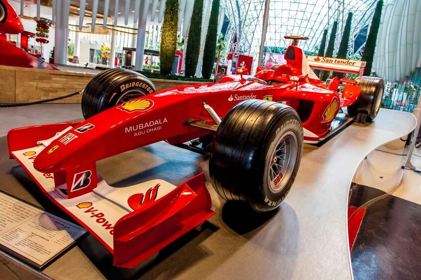 A red Formula 1 car with sponsor logos, displayed indoors with a glass enclosure. 2