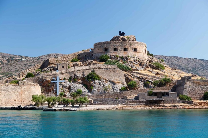 A historic fortress on a hill overlooking a body of water, with a cross and ruins visible. 4