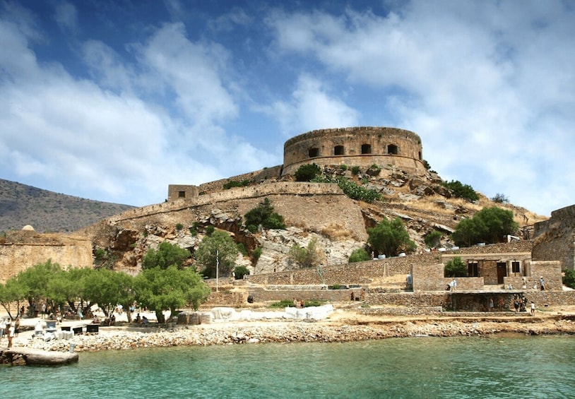 A historical fortress on a hill overlooking a body of water, with ruins and trees in the foreground. 3