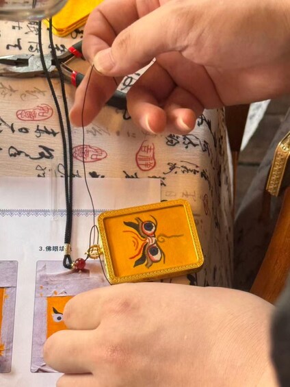 A person holding a small, square, yellow pendant with a design of two eyes and a red and black pattern, attached to a black cord. 3