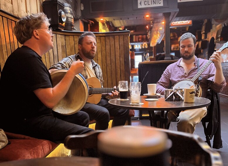 Three people seated at a table with musical instruments and drinks, in a cafe with wooden decor and a sign indicating a clinic. 1