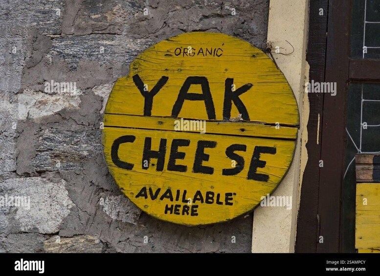A yellow sign with black lettering stating 'YAK CHEESE AVAILABLE HERE' against a stone wall. 2
