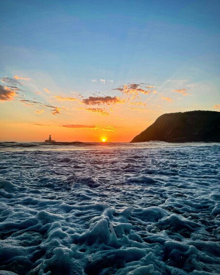 A sunset over a rough sea with a lighthouse in the distance and a cliff on the right. 5