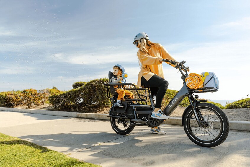 A woman and a child on a tandem electric bike with a basket, riding on a paved path with greenery and a clear sky in the background. 1