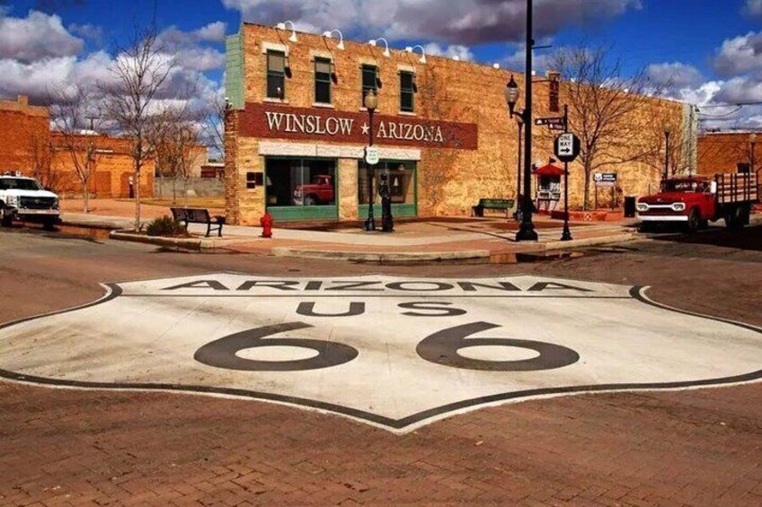 A street with a large ARIZONA 66 sign painted on the road, a building with 'WINSLOW ARIZONA' signage, and a red truck parked on the side. 2